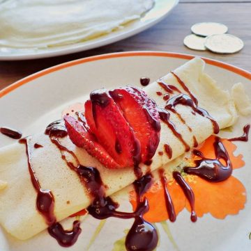 crepe on a plate with chocolate and strawberries on top and text overlay with gold coins and stack of crepes in background.