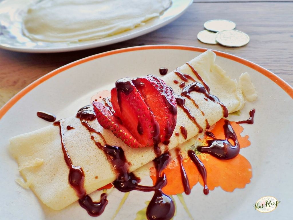 crepe on a plate with chocolate and strawberries on top and text overlay with gold coins and stack of crepes in background.