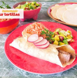 steak burrito on a plate with Mexican rice, avocado salsa and flour tortillas in the background with text overlay "homemade flour tortillas"