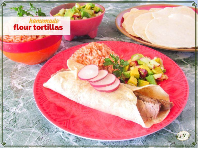 steak burrito on a plate with Mexican rice, avocado salsa and flour tortillas in the background with text overlay "homemade flour tortillas"