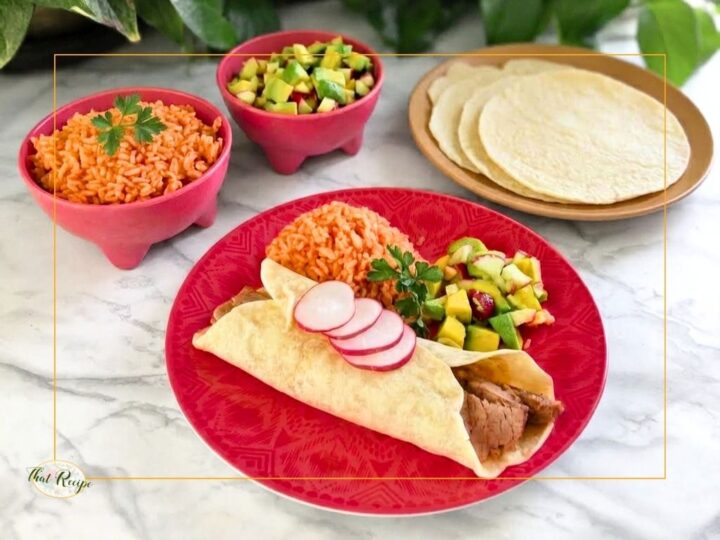 steak burrito on a plate with Mexican rice, avocado salsa and flour tortillas in the background