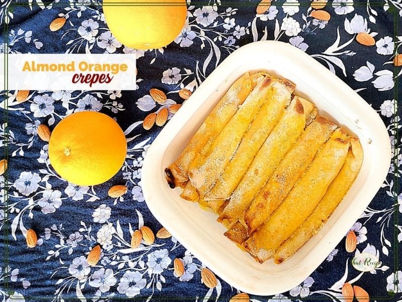 top down view of crepes in a dish surrounded by oranges and almonds with text overlay "almond orange crepes"