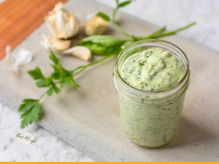 jar filled with green goddess salad dressing on a table with herbs and garlic